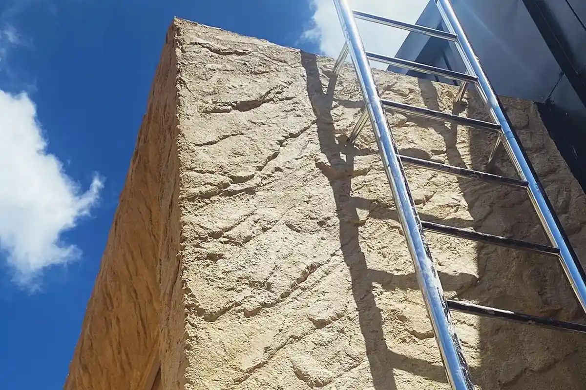 建築外牆轉角的米黃色仿岩飾面，表面有風化岩石般的層狀紋理，旁邊架著不鏽鋼梯